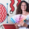 A smiling woman holds a color swatch fan. Behind her is digital art with wavy lines, geometric shapes, and Adobe Illustrator interface elements, hinting at creative courses online for aspiring designers.
