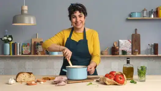 A smiling woman in a yellow top and blue apron stirs a pot in a kitchen, possibly following courses online. On the counter are bread, eggs, garlic, tomatoes, basil, and olive oil. Shelves with books and kitchenware line the background.