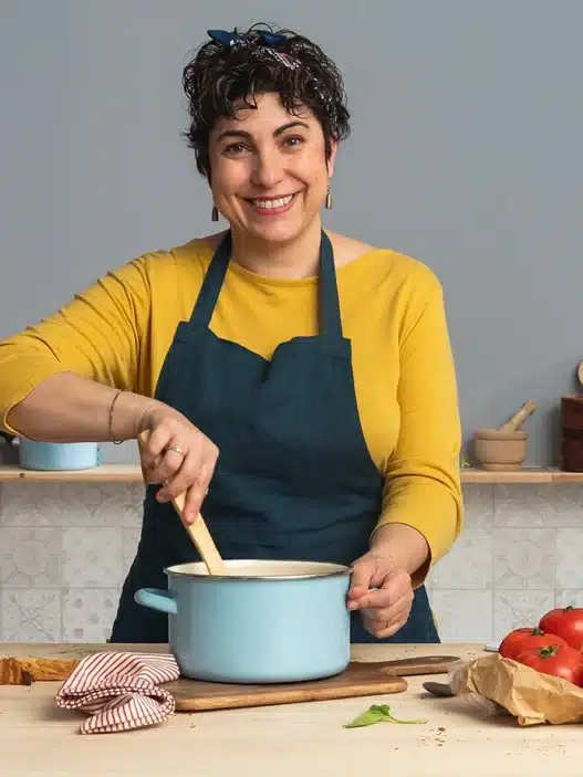 A smiling woman in a yellow top and blue apron stirs a pot in a kitchen, possibly following courses online. On the counter are bread, eggs, garlic, tomatoes, basil, and olive oil. Shelves with books and kitchenware line the background.