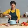 A smiling woman in a yellow top and blue apron stirs a pot in a kitchen, possibly following courses online. On the counter are bread, eggs, garlic, tomatoes, basil, and olive oil. Shelves with books and kitchenware line the background.