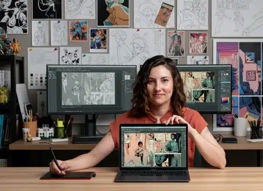 A woman sits at a desk in a studio, holding a pen tablet and displaying a laptop screen with comic art. Behind her are two monitors with similar art and a wall covered in comic sketches and illustrations.