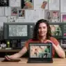 A woman sits at a desk in a studio, holding a pen tablet and displaying a laptop screen with comic art. Behind her are two monitors with similar art and a wall covered in comic sketches and illustrations.