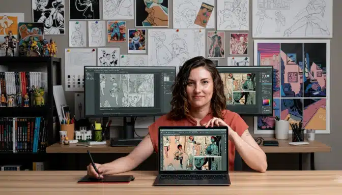 A woman sits at a desk in a studio, holding a pen tablet and displaying a laptop screen with comic art. Behind her are two monitors with similar art and a wall covered in comic sketches and illustrations.