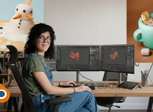 A woman sits at a desk with dual monitors displaying 3D modeling software. On the wall are two 3D art pieces: a melting snowman and a cup of hot chocolate with marshmallows. The Blender logo is in the corner.