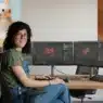 A woman sits at a desk with dual monitors displaying 3D modeling software. On the wall are two 3D art pieces: a melting snowman and a cup of hot chocolate with marshmallows. The Blender logo is in the corner.