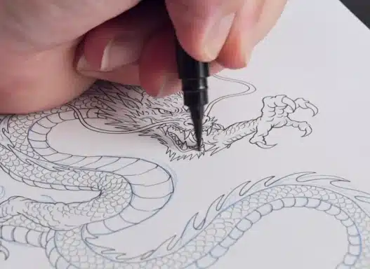 A close-up of a hand using a black pen to outline the head of a detailed dragon drawing on white paper.