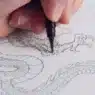 A close-up of a hand using a black pen to outline the head of a detailed dragon drawing on white paper.