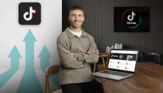 A smiling man sits at a desk with a laptop displaying TikTok on the screen. Behind him, a large TikTok logo and upward-pointing arrows are visible, suggesting growth or success on the platform.