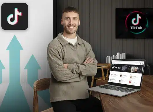 A smiling man sits at a desk with a laptop displaying TikTok on the screen. Behind him, a large TikTok logo and upward-pointing arrows are visible, suggesting growth or success on the platform.