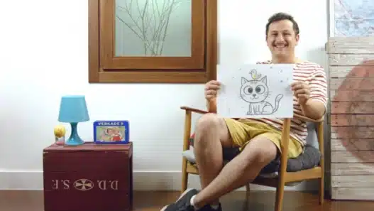 A smiling person sits on a chair in a cozy room, holding up a drawing of a cat with a bird on its head. There is a red box, blue lamp, and a book beside them, and a window and wooden decor in the background.