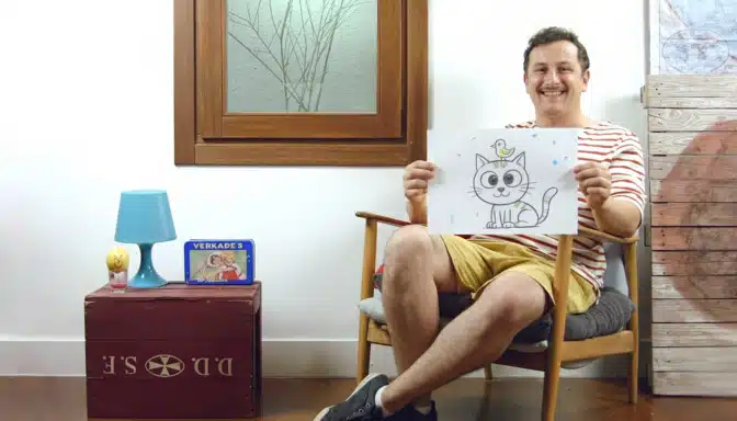 A smiling person sits on a chair in a cozy room, holding up a drawing of a cat with a bird on its head. There is a red box, blue lamp, and a book beside them, and a window and wooden decor in the background.