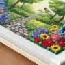 A hand embroiders colorful flowers and a stone path on fabric, with a vibrant garden and a bird in the background, creating a detailed nature scene with thread.