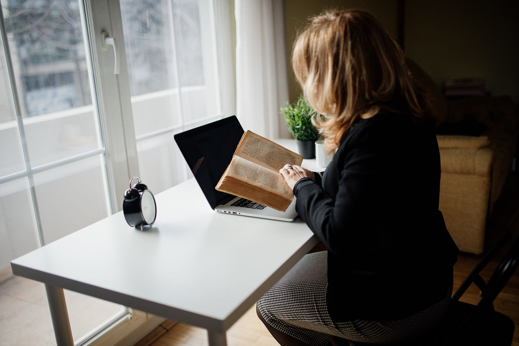 Woman reading a book while using a laptop at home