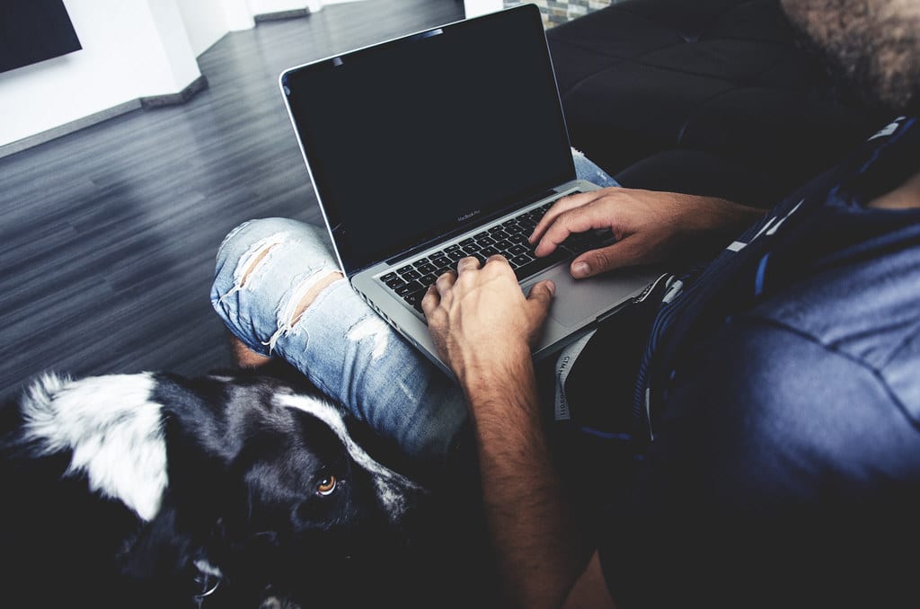 Man typing on a laptop on his lap with a dog beside him