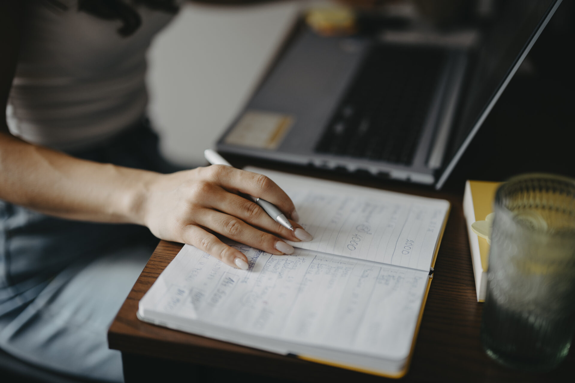 Student writing notes in a notebook while using a laptop at a desk