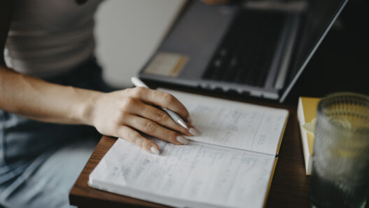 Student writing notes in a notebook while using a laptop at a desk