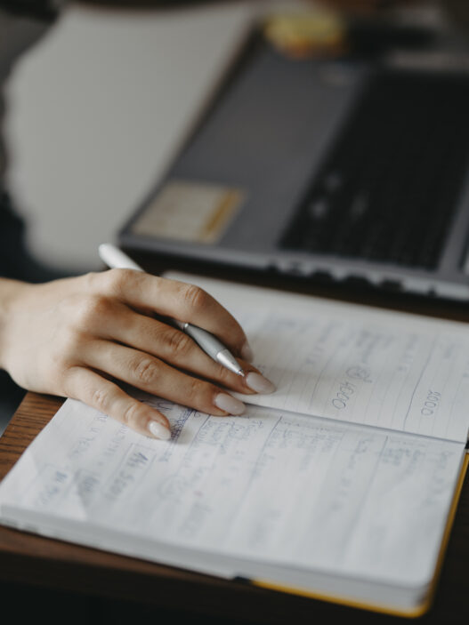 Student writing notes in a notebook while using a laptop at a desk