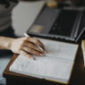 Student writing notes in a notebook while using a laptop at a desk