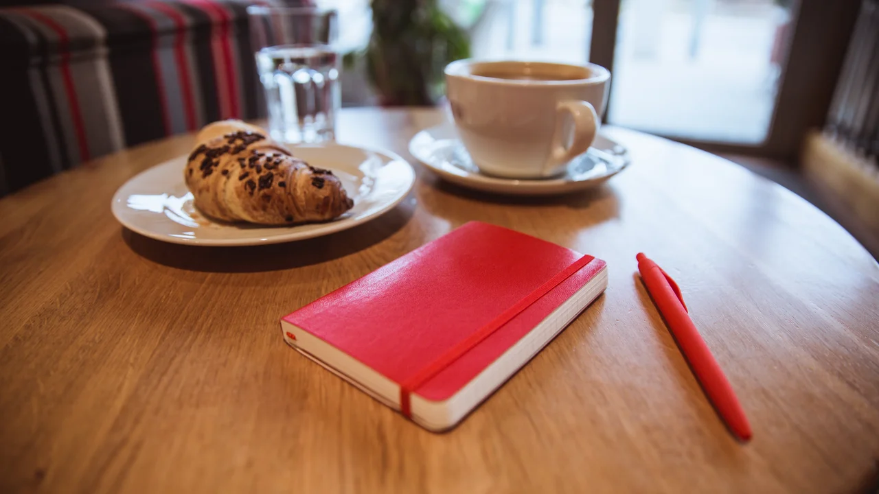 A red note and red pen is on the table in a cafe a cup of coffee and a croissant on background