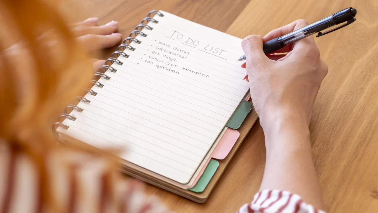 Woman writing a handwritten todo list in a spiral notebook on a wooden desk planning daily tasks and organizing errands productivity and organization in a cozy home workspace