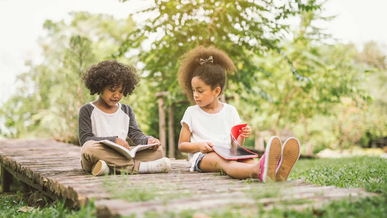 Little child girl reading book between green meadow garden with friend education concept