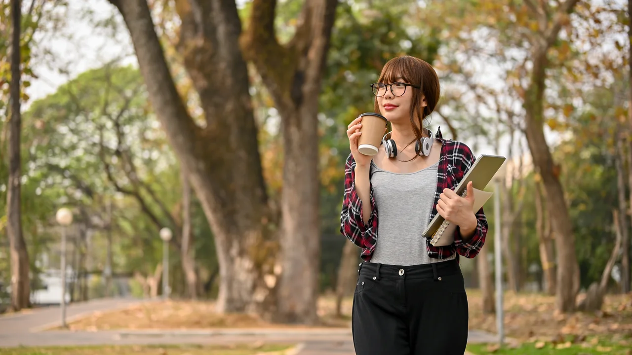 Relaxed asian female college student sipping coffee while walking in the park after finish classes