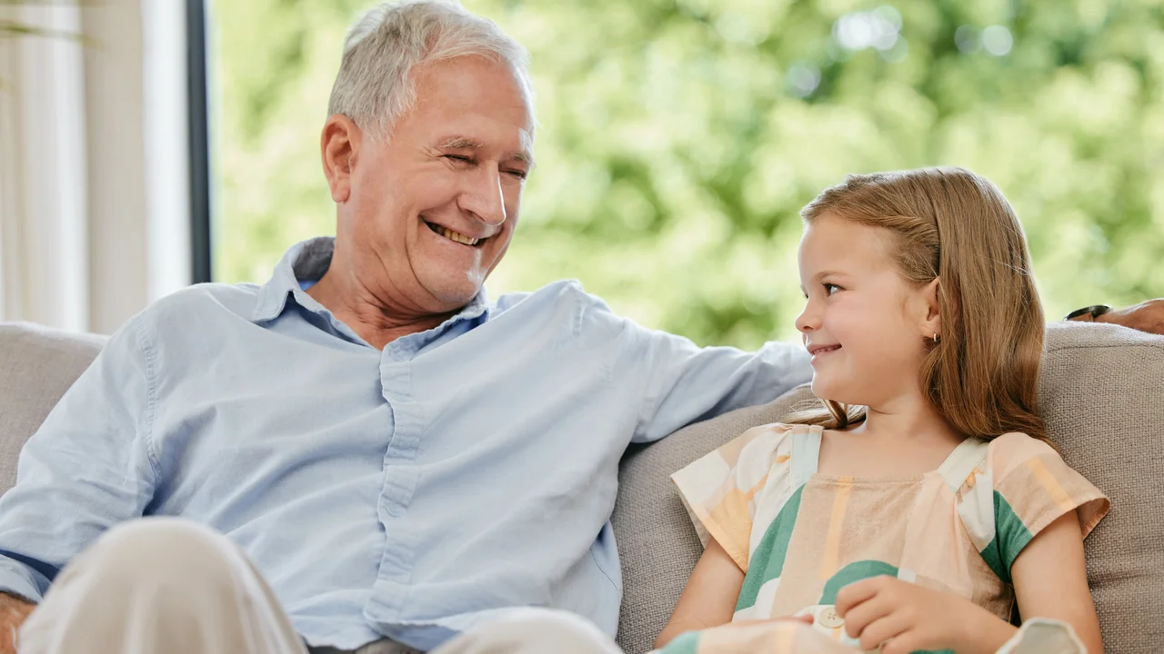 Conversation smile and grandfather with child on a sofa bonding relaxing and talking at home happy family and senior man speaking to young girl kid on a couch in the living room at modern house