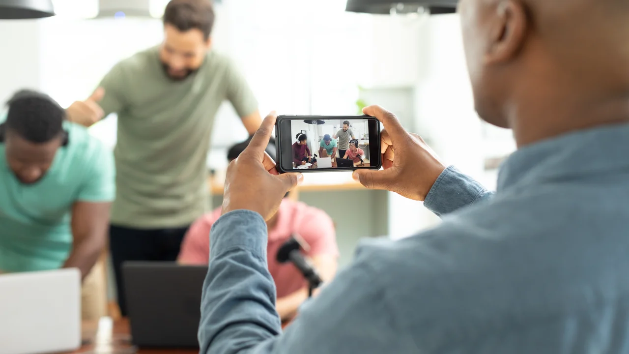Midsection of man filming multiracial male coworkers recording podcast over mobile phone in office. unaltered, interview, broadcasting, communication, media, teamwork and technology concept.